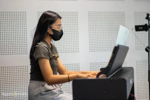Young female pianist with mask playing digital piano with sheet music in soundproofed studio at Legato Music School Kota Kinabalu