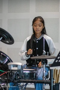 Young female drummer in varsity jacket practicing electronic drums in soundproofed studio at Legato Music