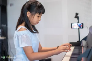 Young girl in blue top practicing piano at home with phone recording setup from Legato Music lessons
