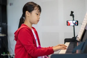 Young girl in red hoodie playing piano with sheet music during online lesson recording from Legato Music School Kota Kinabalu